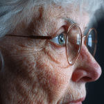 Close-up profile of an elderly woman with glasses, highlighting detailed wrinkles and facial texture, who's suffering from macular degeneration challenges