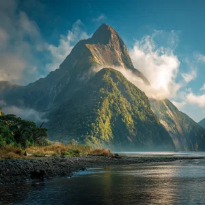 Mitre Peak reflecting in the calm waters of Milford Sound at sunrise, with mist and lush green cliffs in the background.
